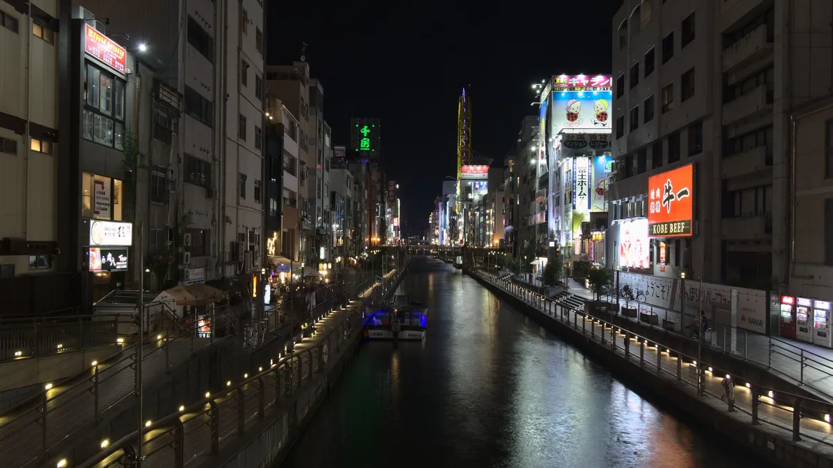A nightscape of the retail and entertainment district. A straight channel runs through the center of the image. Pedestrian pathways are on the left and right of the river, both down at the water and a bit raised, to give access to shops and restaurants. Ramps allow passing between the different levels and bridges in the distance close the gap between both shores while allowing passenger boats to pass under. The varicolored and lit signs reflect in the water of the river.