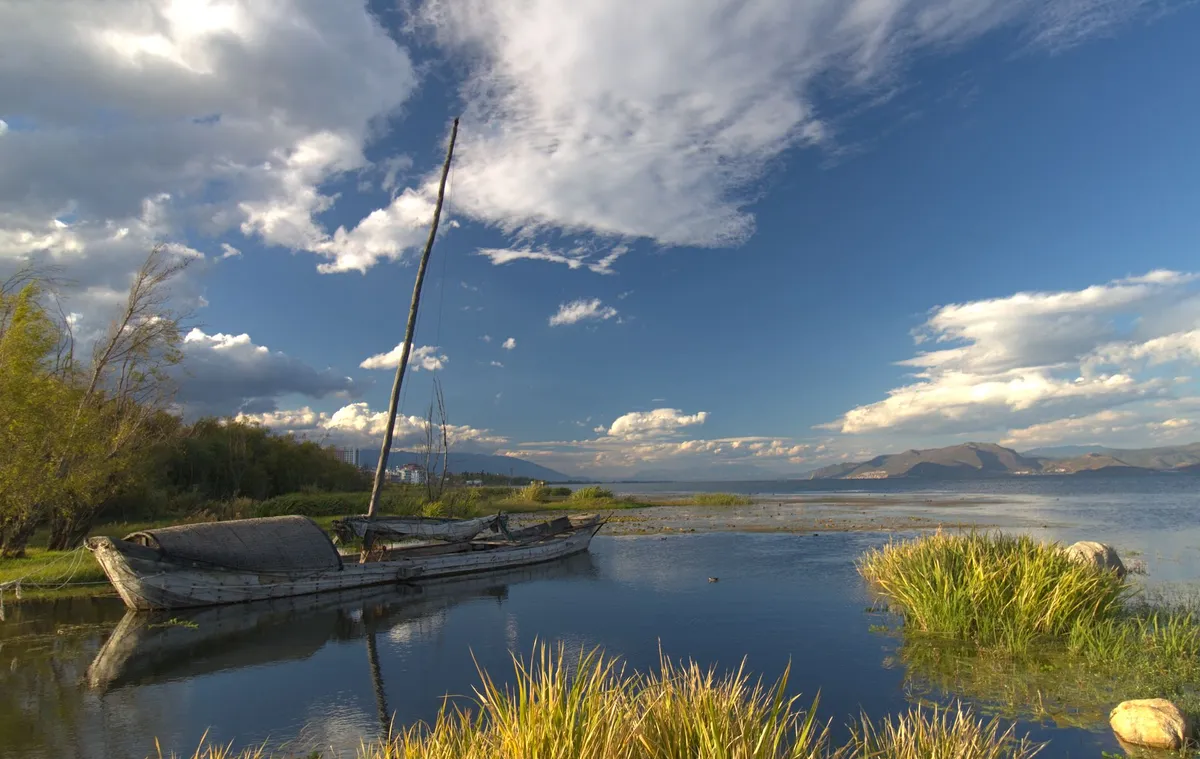A vast lake stretches out to the horizon. Mountains are visible far behind the lake. The water is as deep blue as the sky, as it reflects its color. Yet various clouds cover part of the sky.
An old long boat with an unused mast ran aground in an area of low water level. Behind the boat the shallow water transitions to a swamp.
2 tufts of reed comprise the foreground, with the one on the right reflecting from the surface of the water. 2 rocks protrude through the water surface in the lower right, behind and in front of the reed.