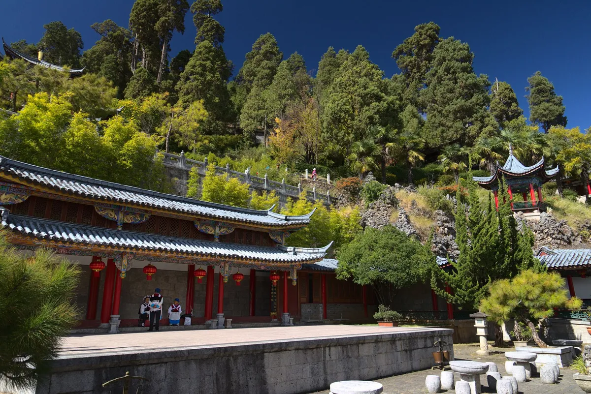A roof system in traditional chinese style is marking the end of a park with a few stone tables, stools and nicely cut Trees. Under this roof are three locals in traditional clothing. Behind this roof a hill with dens greenery is expanding upwards. A path is making its way up the hill from the right with a few pavilions along the way up.
