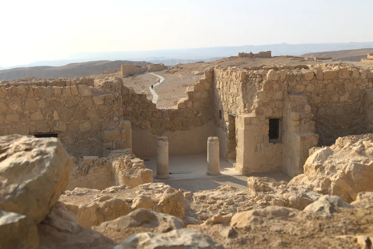 Ancient looking ruins dominate the scene, with two pillars in the center. In the background there is a path making its way to another ruin in the distance, with two people walking there. All stones and the ground have a yellow-ish sandstone like hue.