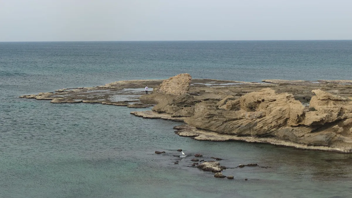 A view of the Ocean. A rock shelf is pretruding into the image from the right side with a single person standing on there gazing into the distance.