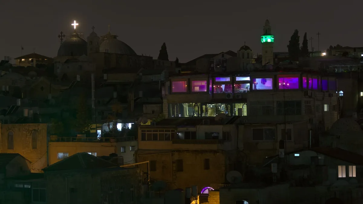 A night view of Jerusalem. In the right seems to be a night club, because of its colorful purple lighting. In contrast in the left is a church with a white lit christian cross.